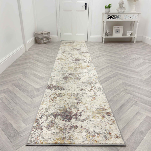 The Couture 52023-6616 Runner Rug, featuring a mottled abstract pattern in neutral tones, is placed on a light herringbone wood floor in a bright hallway with white walls, a white door, and a small white console table.