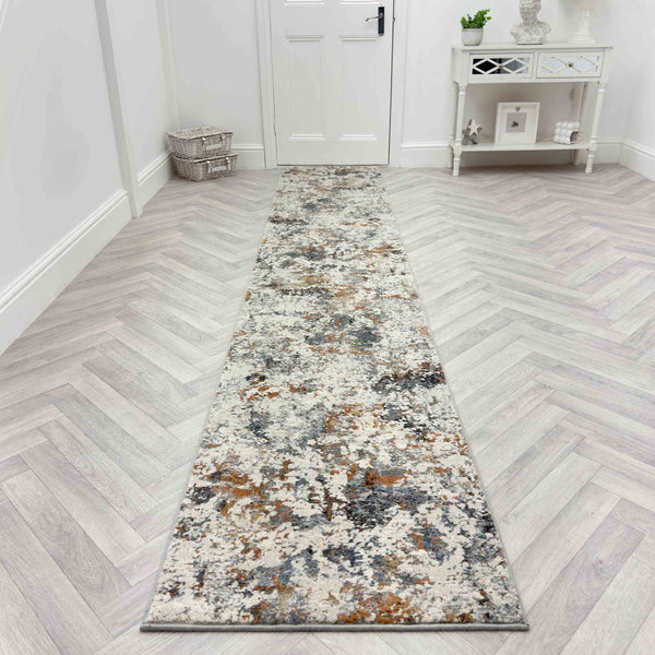 The Couture 52023-6616 Runner Rug, featuring a mottled gray, beige, and brown pattern, lies on a light herringbone wood hallway floor with a white door, console table, and decor in the background.
