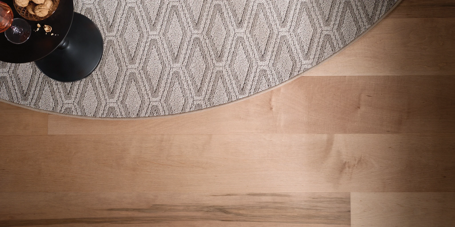A round, patterned gray rug is partially shown over a light wood floor. A small black table with a glass of pink liquid and snacks is visible in the upper left corner.