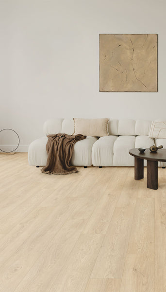A modern living room with a white, plush sofa, a beige throw blanket, a light pillow, a round dark wooden coffee table, minimalist decor, and a light wood floor. A large abstract painting hangs on the wall above the sofa.