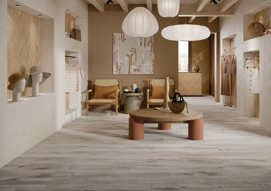 A stylish boutique with neutral tones features wooden floors, modern pendant lights, woven chairs, a round coffee table, wall art, clothes on display, and hats on mannequin heads in wall niches.