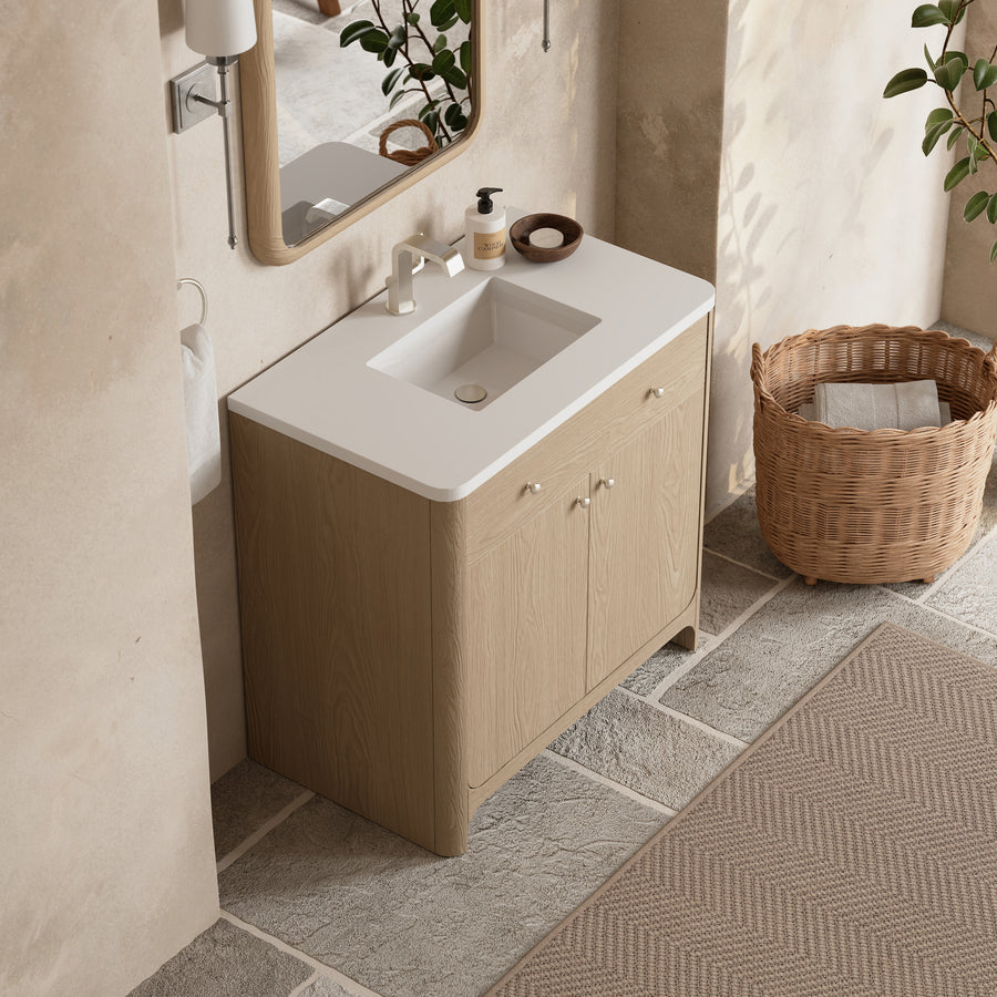 A modern bathroom vanity with a light wood cabinet, white countertop, and rectangular sink, set against beige stone walls. A mirror, soap dispenser, and a wicker basket are nearby, with green plants in the background.