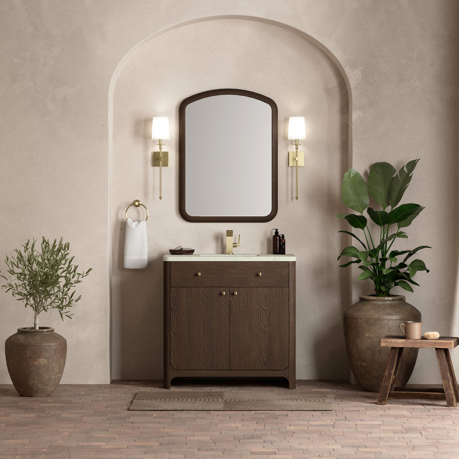 A stylish bathroom features the Gracyn 36" Vanity in dark wood, a large arched mirror, two wall sconces, a towel ring, a potted olive tree, and a leafy plant in a rustic pot against a neutral textured wall for transitional appeal.