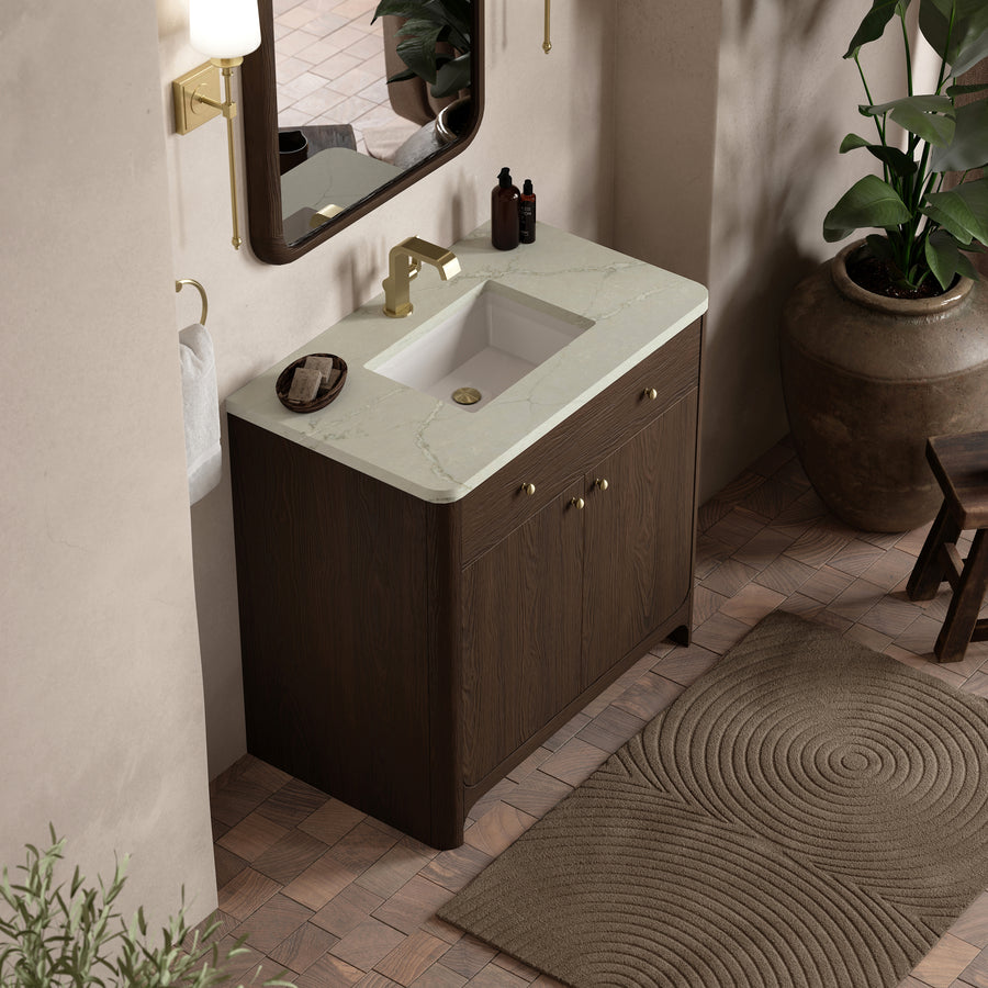 A modern bathroom vanity with a marble countertop, gold faucet, wood cabinet, and rectangular mirror. The space features a beige wall, potted plants, a brown textured rug, and a towel hanging on a round holder.
