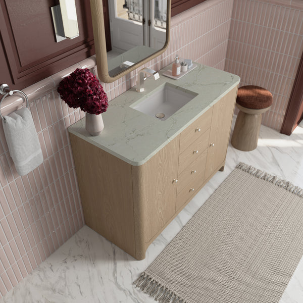 Modern bathroom with a light wood vanity, marble countertop, sink, and faucet. Red flowers in a vase, soap dispenser, and towel on the side. Pink tile wall, a rug, a wooden stool, and a large mirror above.