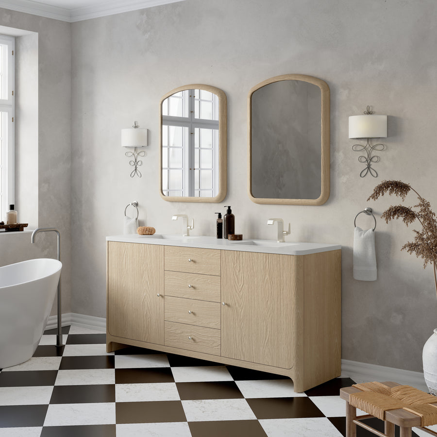 Modern bathroom with a light wood vanity, two rectangular mirrors, wall-mounted lights, a white freestanding bathtub, and a black-and-white checkered tile floor. Dried plants decorate a wooden stool in the corner.