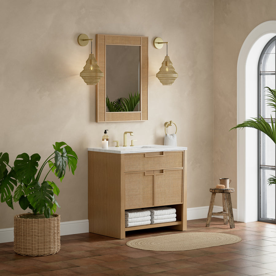 A modern bathroom with beige walls, a wooden vanity with a white countertop, a rectangular mirror, gold fixtures, pendant lights, a round rug, a potted plant, a wooden stool, and an arched window with natural light.
