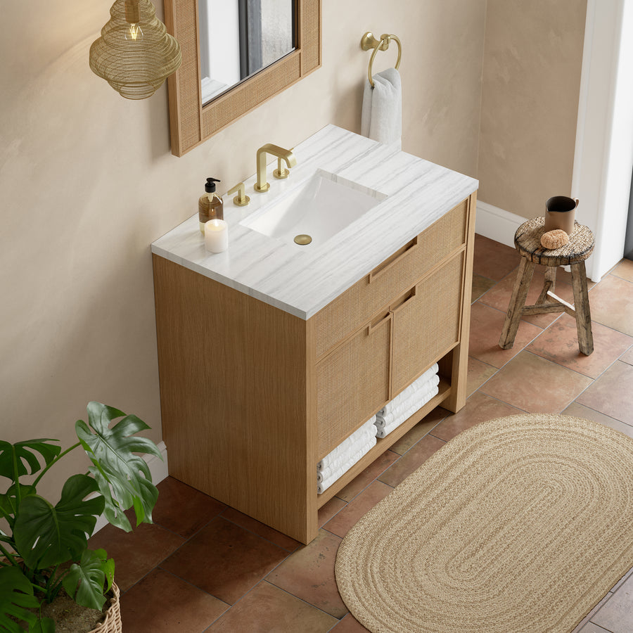 A modern bathroom with a wooden vanity, white marble countertop, gold faucet, soap dispenser, hand towel, rolled towels below, potted plant, woven rug, stool, and terracotta floor tiles.