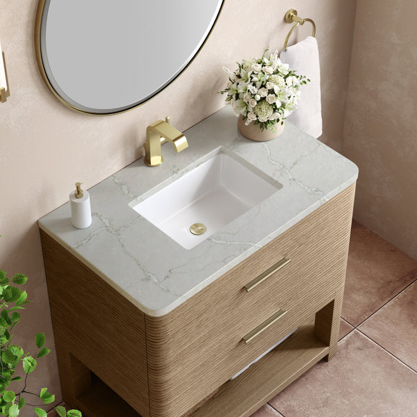 A modern bathroom vanity with a marble countertop, gold faucet, soap dispenser, potted white flowers, and a towel on a gold ring. A round mirror hangs above, and a green plant is visible in the corner.