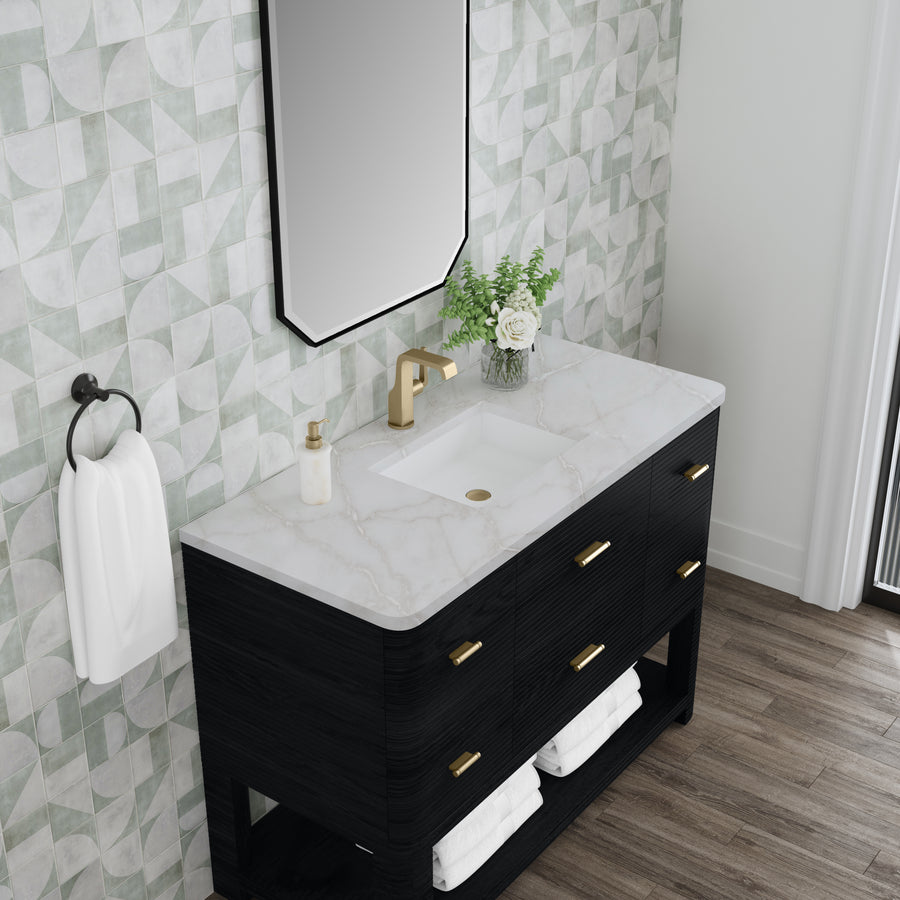 A modern bathroom vanity with a marble countertop, gold faucet, and black cabinet with gold handles. A rectangular mirror, vase with white flowers, soap dispenser, towel ring with a white towel, and geometric wall tiles are visible.