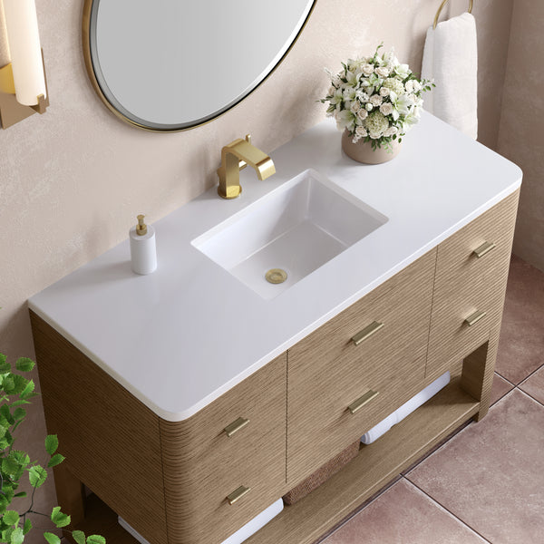 A modern bathroom vanity with a white countertop, rectangular sink, gold faucet, soap dispenser, and a potted white flower arrangement. A round mirror and towel ring are mounted on a beige wall above.