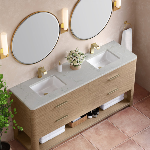 A modern bathroom with a double vanity featuring two sinks, oval mirrors, gold faucets and lights, a vase of white flowers, rolled towels, and shelves with toiletries. The floor has terracotta tiles and there’s a green plant nearby.