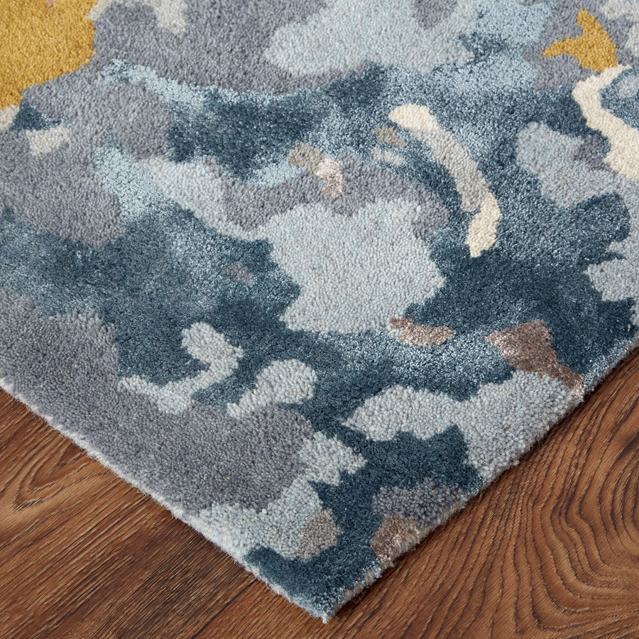 A close-up of a rug with an abstract blue, gray, and beige pattern, placed on a wooden floor. The corner of the rug is visible, showing textured, multicolored design details.