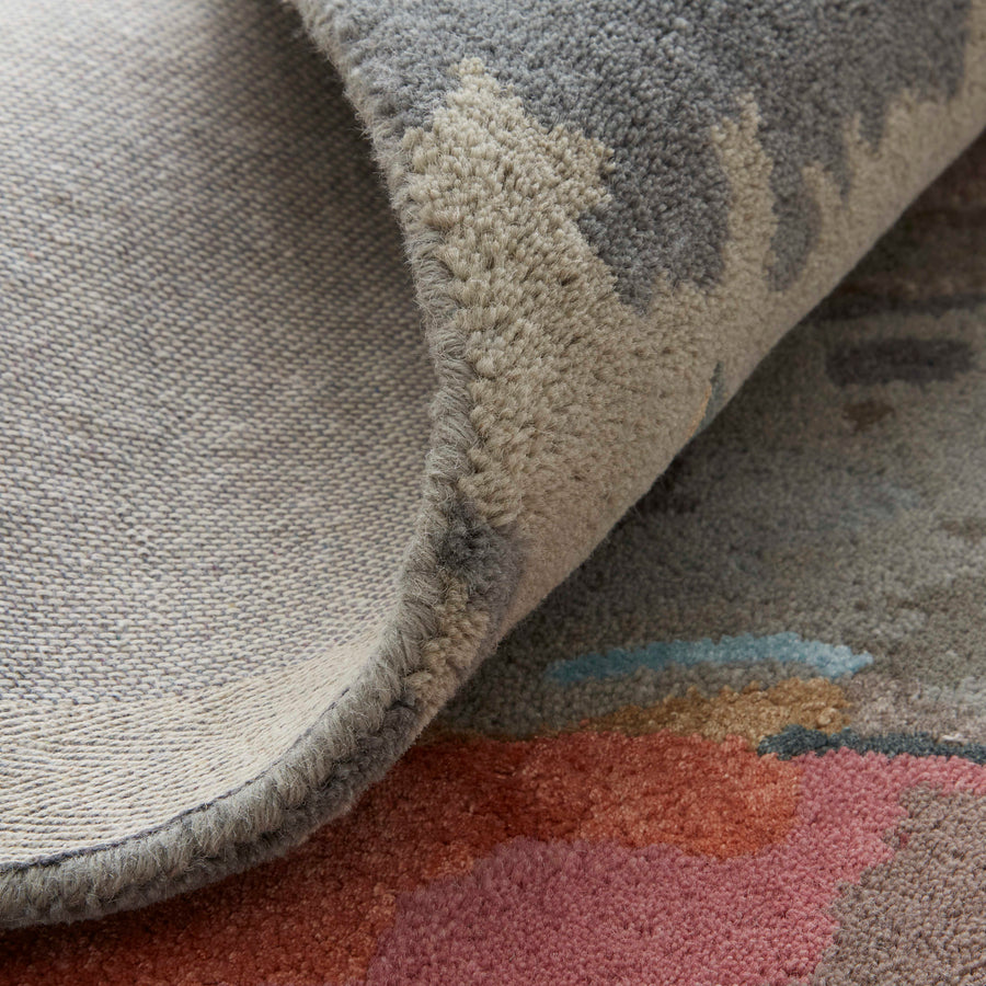 A close-up of a rugs corner folded over, showing its textured top side with abstract patterns and the flat, woven underside. The pattern includes colors like gray, beige, red, and blue.