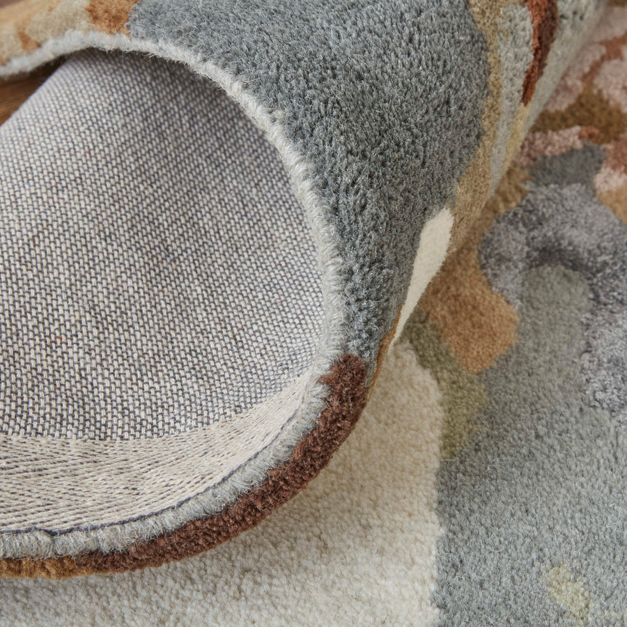 A close-up of a corner of a multicolored, textured rug being lifted to show the fabric backing and stitched edge detail.
