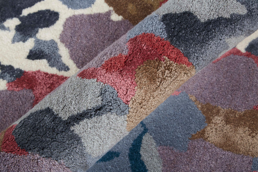 Close-up of a textured, multicolored rug with abstract patterns in shades of purple, red, gray, brown, and cream. The rug is partially rolled, showing its plush surface and intricate design.