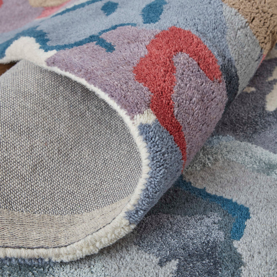 A close-up of a colorful rug corner folded over, showing the textured top with abstract blue, purple, and red patterns, and the gray woven underside.