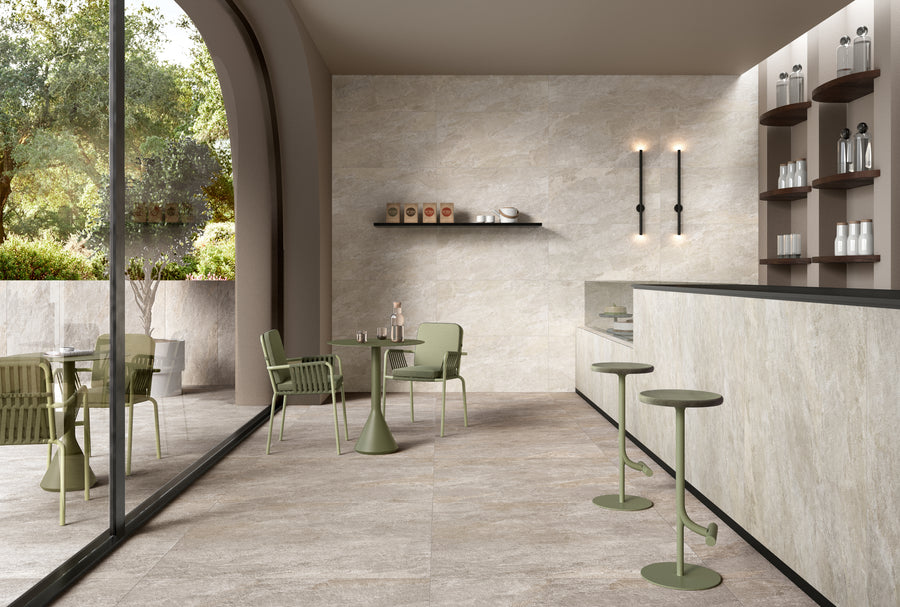 Modern cafe interior with beige stone walls and floor, green chairs and tables, bar area with shelves of cups, and large window overlooking a garden with trees. Natural light fills the stylish, minimalist space.