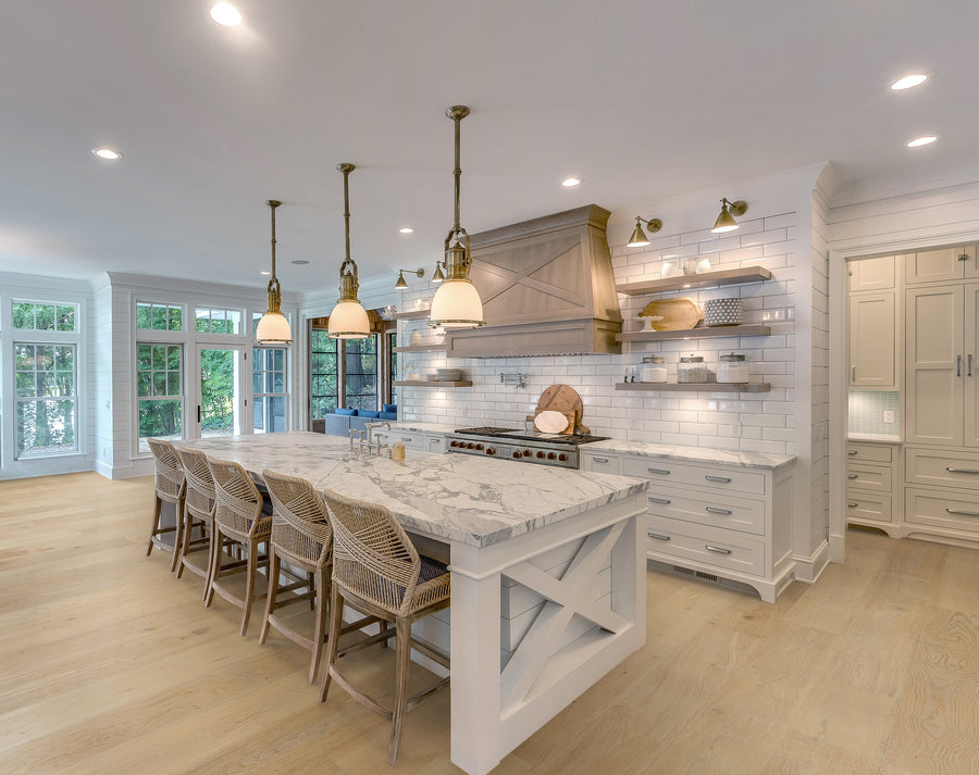 Spacious modern kitchen with a large marble island, wicker bar stools, pendant lights, white cabinets, open shelves, stainless steel stove, and wood floors. Large windows provide natural light.
