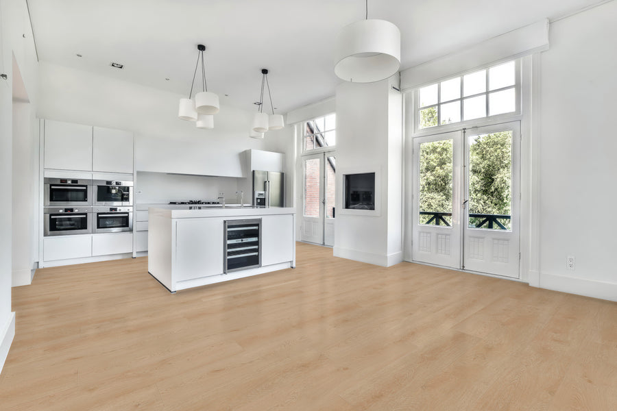 A modern, bright kitchen with white cabinets and island, built-in appliances, light wood flooring, pendant lights, large windows, and French doors opening to a green view.