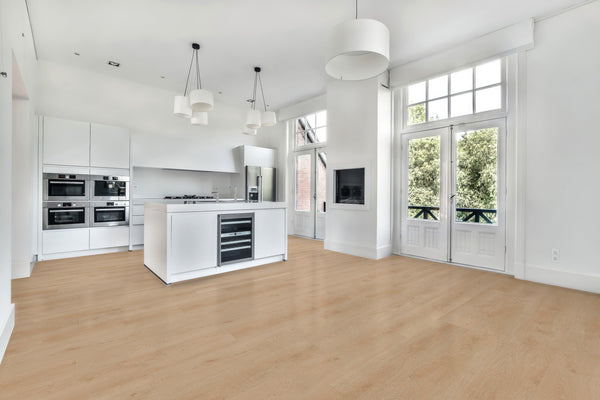 A modern, bright kitchen with white cabinets and island, built-in appliances, light wood flooring, pendant lights, large windows, and French doors opening to a green view.