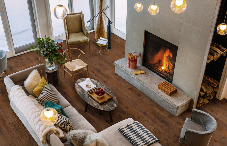 A cozy living room with a lit fireplace, modern furniture, throw pillows, a wood coffee table with decor, a vase of greenery, and large windows letting in natural light. Firewood is stacked beside the fireplace.
