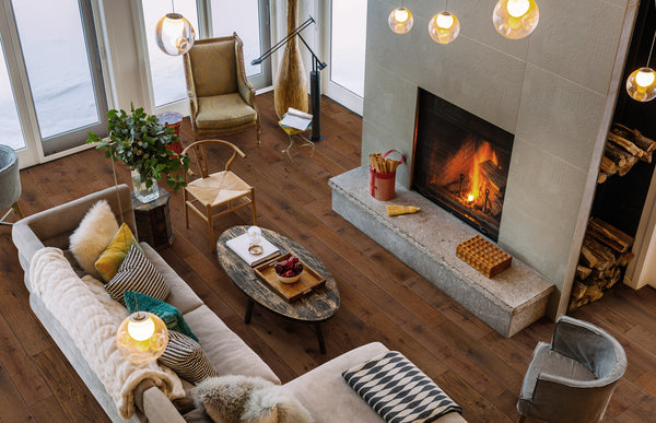 A cozy living room with a lit fireplace, modern furniture, throw pillows, a wood coffee table with decor, a vase of greenery, and large windows letting in natural light. Firewood is stacked beside the fireplace.