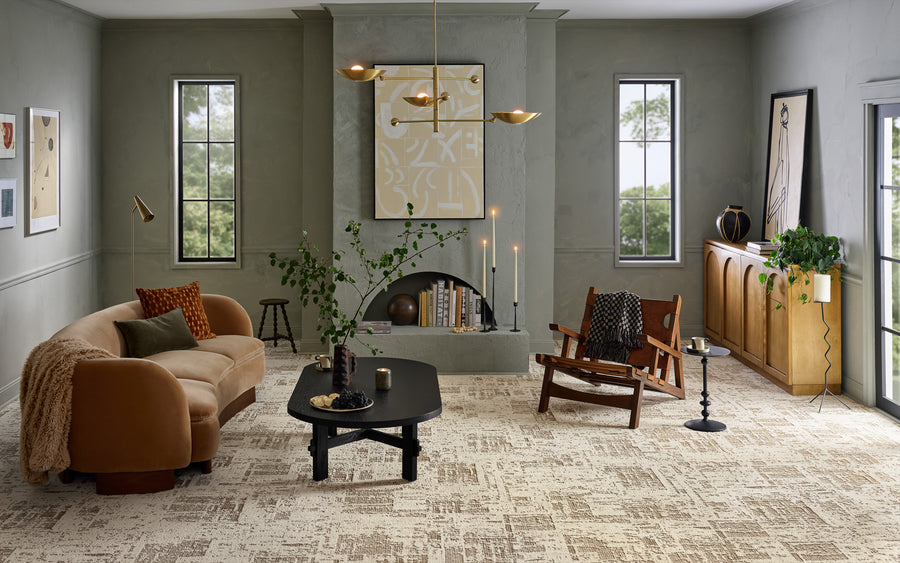 A modern living room with light gray walls, large windows, a curved beige sofa, black coffee table, wooden armchair, green plants, abstract art above a fireplace, and a wooden sideboard against the right wall.
