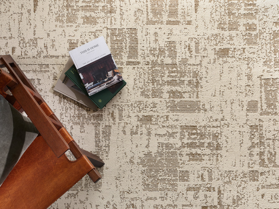 A wooden chair with a gray cushion is next to a stack of books, including one titled Tings by Range, on a beige textured rug with a geometric pattern.