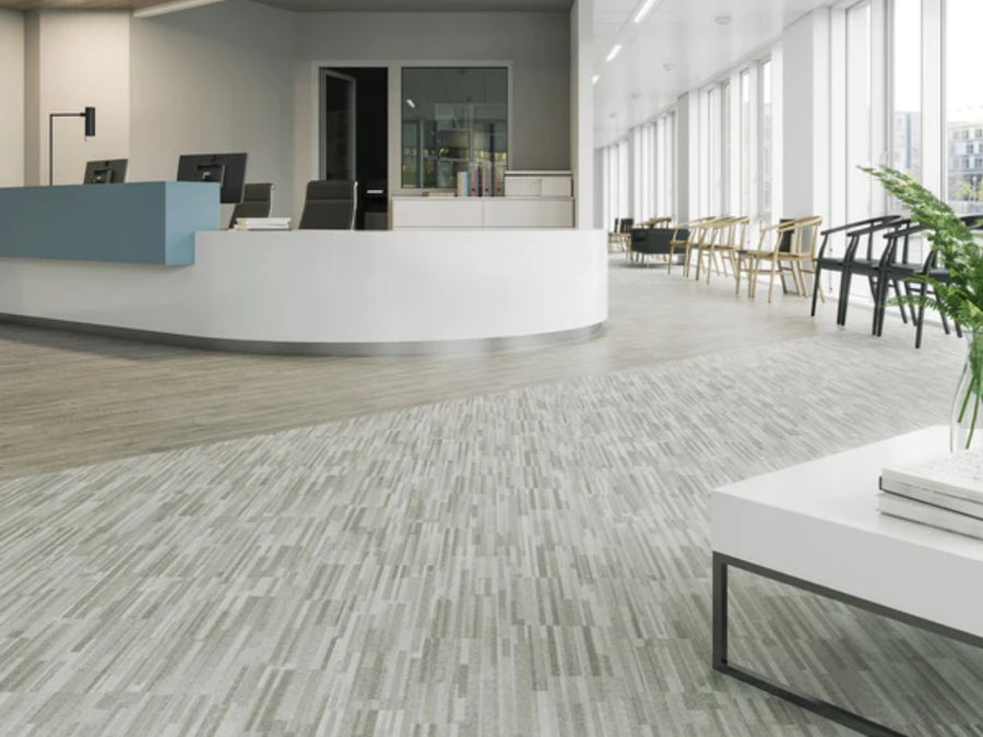 Modern office lobby featuring a curved reception desk, desktop computers, large windows, a row of wooden chairs, and Commercial Vinyl flooring with a white table and green plant in the foreground.