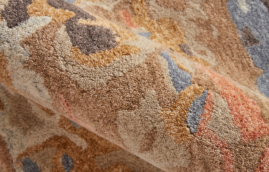 A close-up view of a rolled, textured, multicolored rug with abstract patterns in shades of beige, brown, blue, and orange. The fibers appear soft and plush.