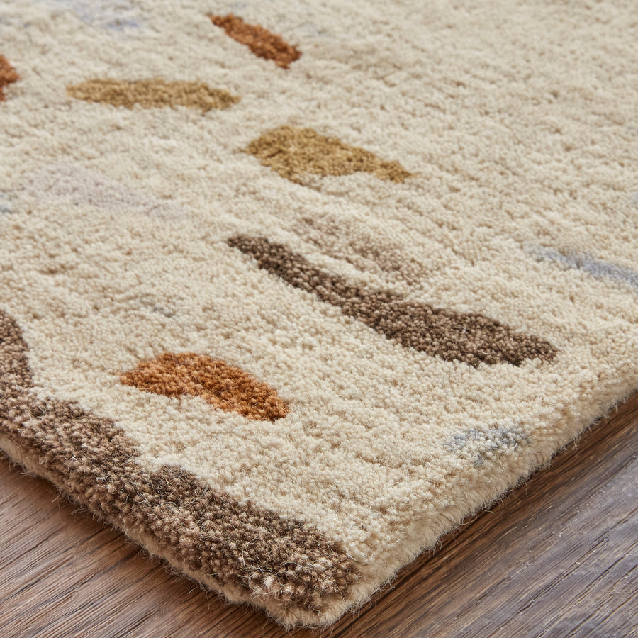 Close-up of a beige textured rug with abstract shapes in brown, tan, and rust tones, placed on a wooden floor. The rug appears thick and soft, with a mix of short and medium pile heights.