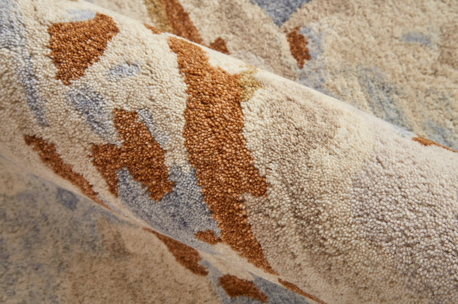 A close-up view of a textured, plush rug with an abstract pattern in beige, brown, and light blue tones. The fibers and soft surface detail are visible.