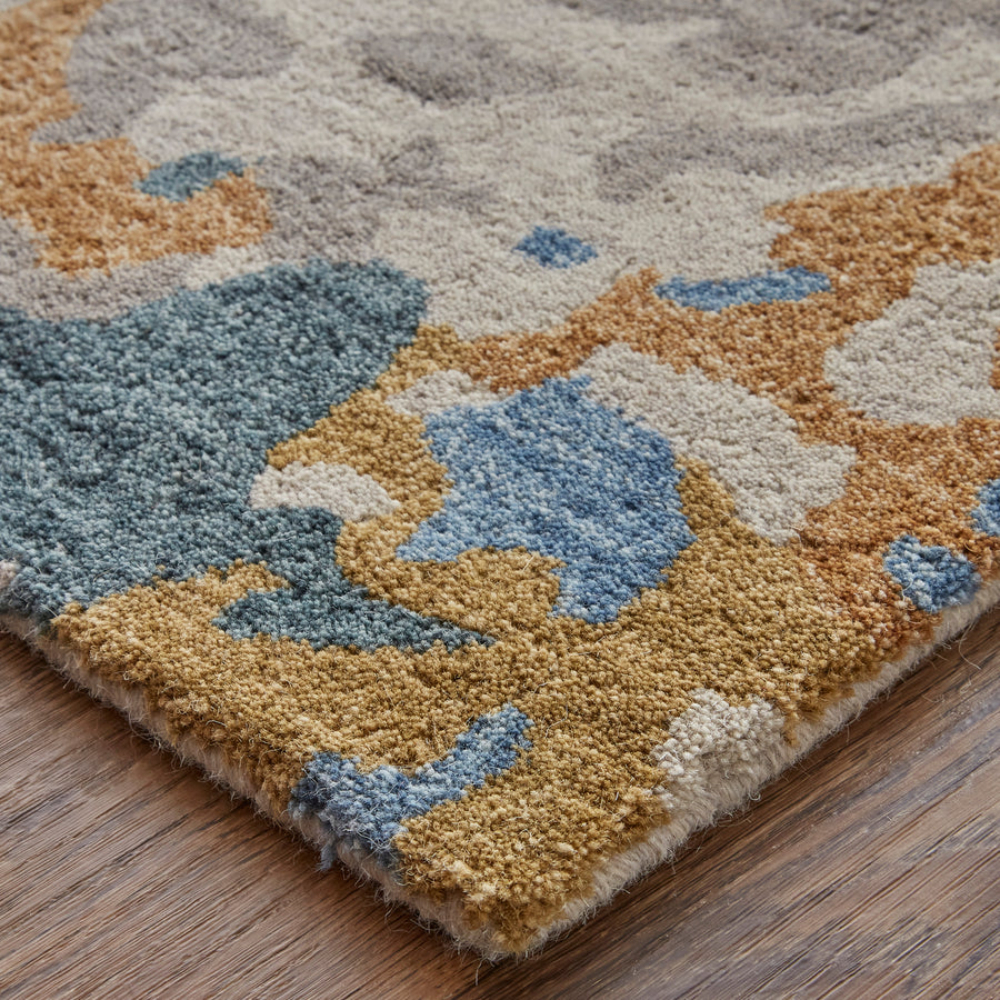 A close-up of a rug with an abstract pattern in blue, beige, gray, and mustard yellow hues on a wooden floor. The textured design features irregular shapes and a soft, tufted surface.