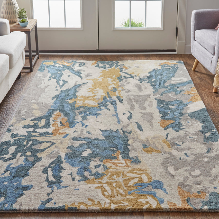 A modern living room with a patterned area rug featuring blue, beige, gold, and gray abstract designs on a wooden floor, surrounded by a sofa, armchair, side table, and large windows with sunlight.