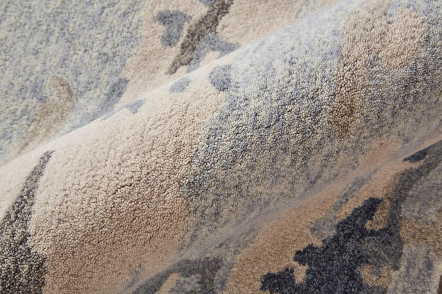 Close-up of a textured, patterned carpet with beige, gray, and blue tones. The carpet appears folded, showcasing its soft, plush surface and abstract design.