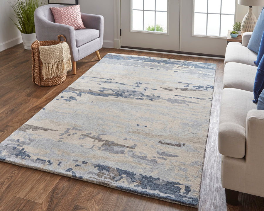 Modern living room with a gray armchair, beige sofa, and a large area rug featuring blue and beige abstract patterns, set on wood flooring near double glass doors with sunlight streaming in.