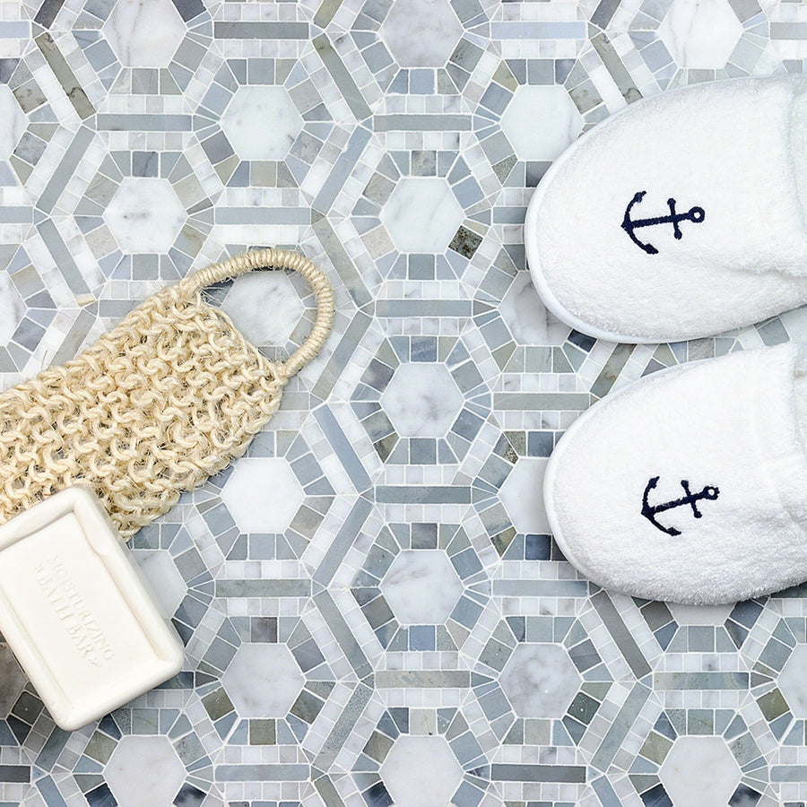 White slippers with blue anchors, a textured loofah, and a bar of soap are arranged on a geometric, gray-and-white tiled floor.