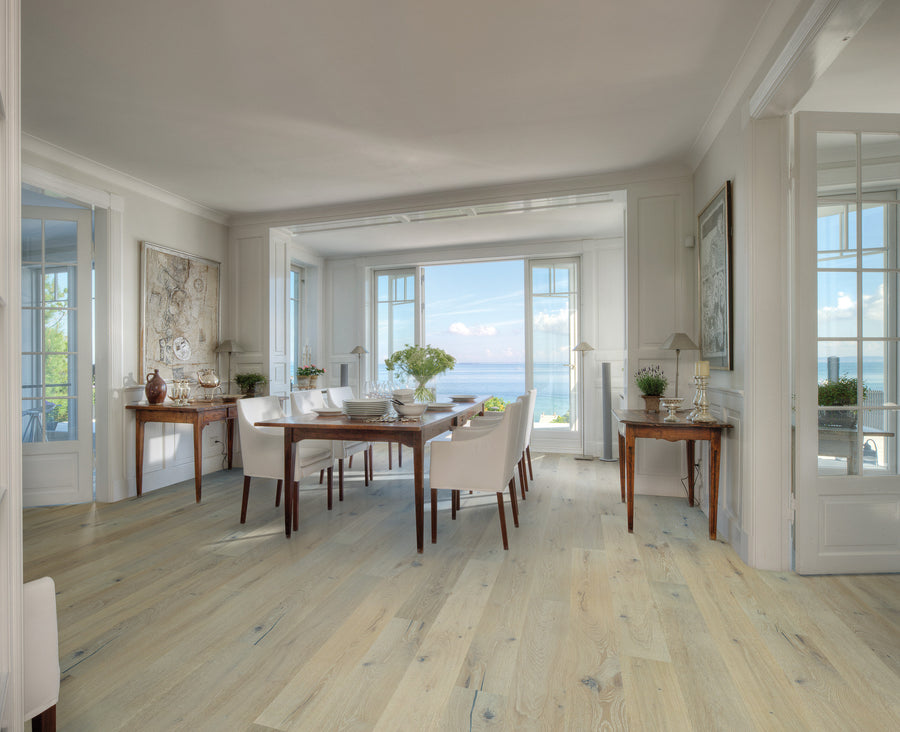 Bright dining room with light wood floors, white walls, and large windows overlooking the ocean. A wooden dining table with white chairs sits in the center, surrounded by elegant decor and plants.