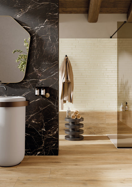 Modern bathroom with a black marble wall, round mirror, white sink, towels hanging on a brick-style beige wall, wooden floors, and a small stand holding rolled towels and toiletries.