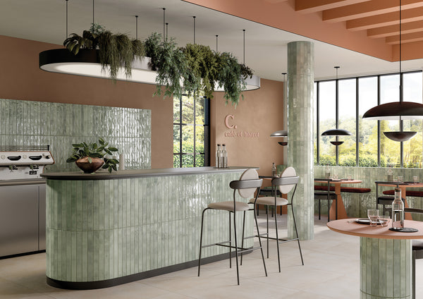 Modern café interior with glossy green tile counters, tall chairs, a hanging planter with greenery, large windows letting in natural light, and tables set for dining. Warm brown accents and sleek lighting create a cozy atmosphere.