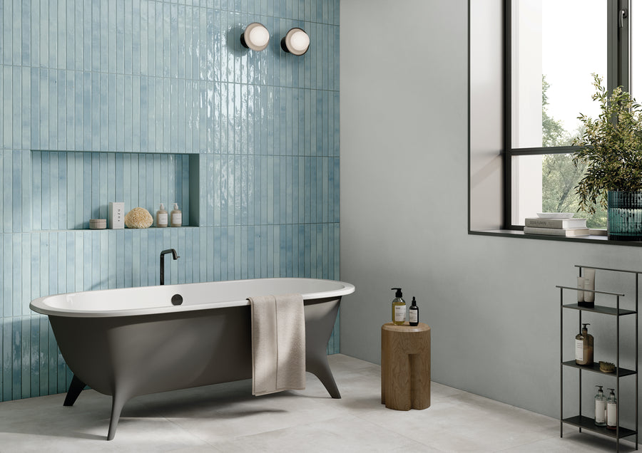 Modern bathroom with a freestanding gray bathtub, blue tiled wall, towel draped over tub, wooden stool with toiletries, shelf with products, and a large window letting in natural light.