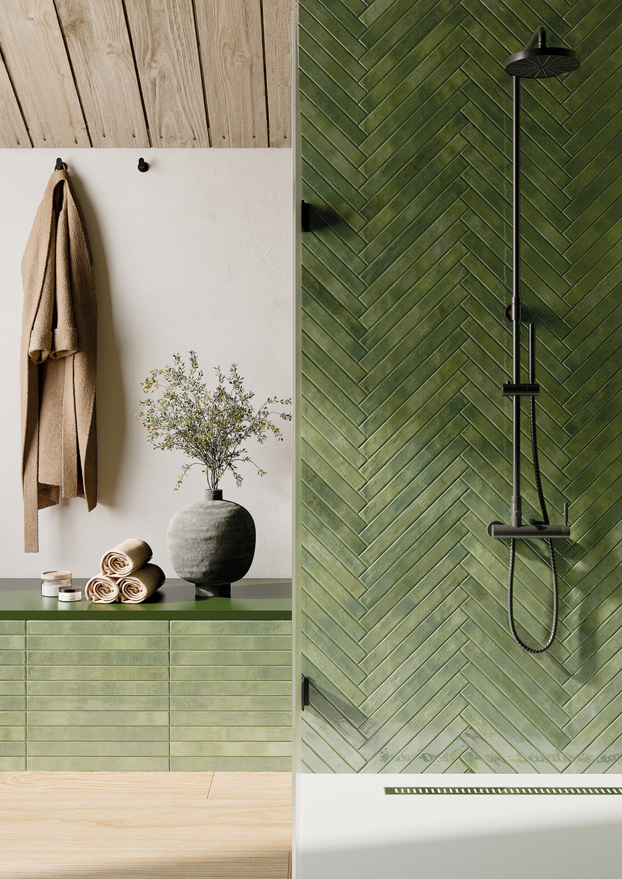 A modern bathroom with green herringbone tile shower, black shower fixtures, olive green vanity with a vase of flowers, rolled towels, and a beige towel hanging on the wall.