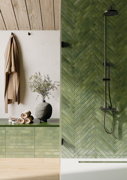A modern bathroom with green herringbone tile shower, black shower fixtures, olive green vanity with a vase of flowers, rolled towels, and a beige towel hanging on the wall.