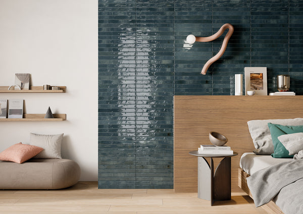 Modern living space with glossy dark teal tiled wall, light wood furniture, a gray ottoman with pillows, floating shelves with decor, and a sculptural wall lamp above a minimalist bed. Light and neutral tones dominate the room.