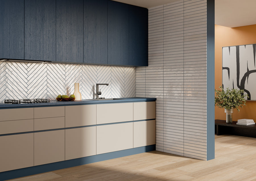 Modern kitchen with blue upper cabinets, beige lower cabinets, and a herringbone-patterned backsplash. A sink and a gas stovetop are on the counter. Wall art and a plant are visible in the adjacent orange-walled room.