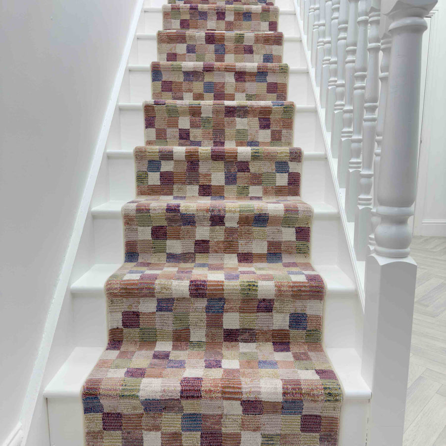 A white staircase features the Eclipse 63339-6121 Runner Rug, showcasing small multicolored squares in beige, brown, pink, and purple. White railings with spindles line the right side.