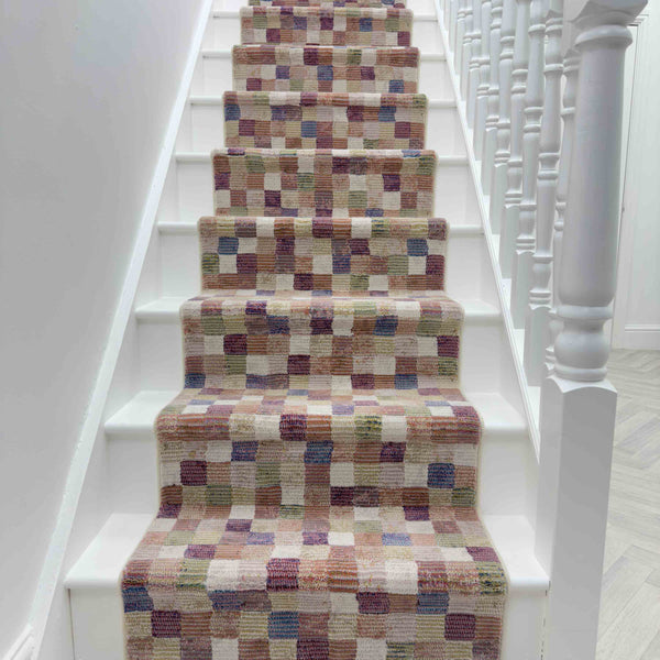 A white staircase features the Eclipse 63339-6121 Runner Rug, showcasing small multicolored squares in beige, brown, pink, and purple. White railings with spindles line the right side.