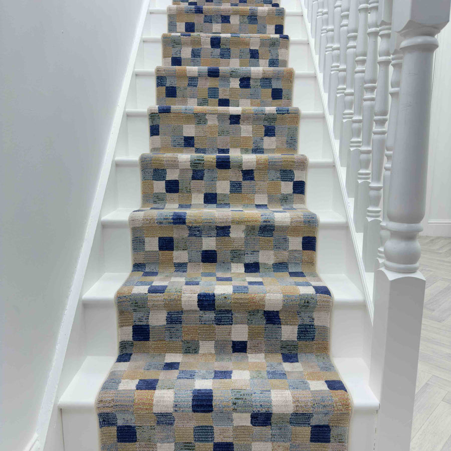 A white staircase with the Eclipse 63339-6121 Runner Rug, displaying blue, beige, and gray squares and rectangles, is flanked by white railings on the right.