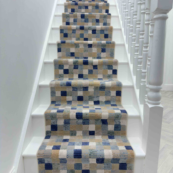 A white staircase with the Eclipse 63339-6121 Runner Rug, displaying blue, beige, and gray squares and rectangles, is flanked by white railings on the right.